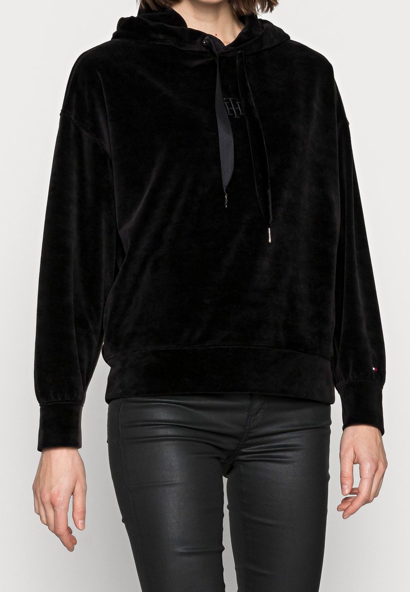 Femme portant un sweat à capuche en velours noir avec cordons et un pantalon en cuir noir ajusté sur un fond uni.