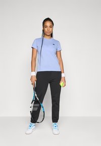 Kvinne i lys blå skjorte, svarte bukser, hvite joggesko som holder en tenniskølle og ball, står foran en enkel lys bakgrunn.