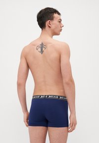 Jūras zila bokseru bikses ar melnu zīmola jostu, kas izceļas ar gludu auduma tekstūru un redzamu tetovējumu, kas veido fleurde-lis.