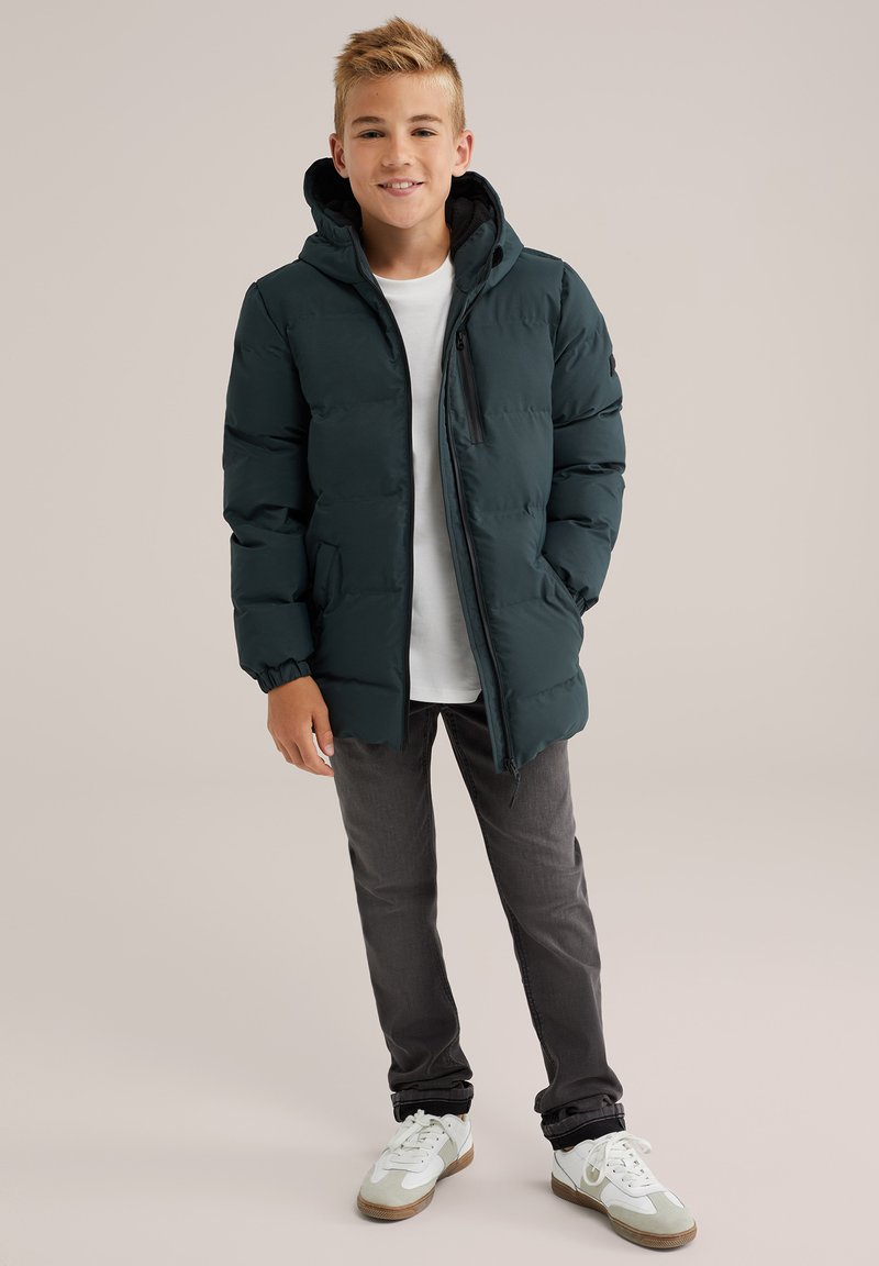 Temno zelen puffer jakna s kapuco, z zadrgo spredaj in elastiko na rokavih, v kombinaciji z sivimi kavbojkami in belimi supergami.