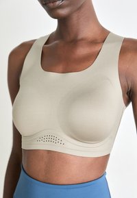 Kvinne iført en beige treningsbh med perforerte detaljer under koppene og høyhvelte blå leggings mot en ensfarget bakgrunn.
