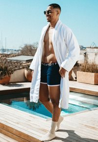 Vit badrock över marinfärgade boxerkalsonger med vit kant. Ljusfärgade strumpor. I bakgrunden finns en pool och krukväxter, under en klar himmel.