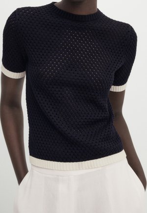 Haut en maille bleu marine foncé à manches courtes avec une bordure blanche aux manches et à l’ourlet, porté avec un pantalon blanc taille haute.