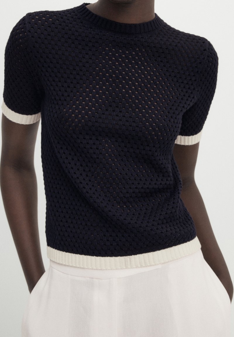 Haut en maille bleu marine foncé à manches courtes avec une bordure blanche aux manches et à l’ourlet, porté avec un pantalon blanc taille haute.