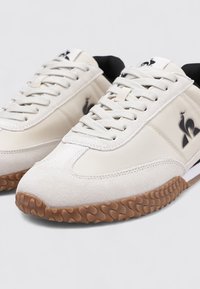 Lichtbeige sneakers van suède en leer, met een gestructureerde rubberen zool, zwarte accenten en een subtiele logodetail aan de zijkant.