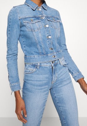 Personne portant une veste en jean bleu clair avec fermeture à boutons et un jean taille haute assorti, debout devant un fond uni.