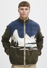 Giacca in pile con colletto alto; design di un paesaggio montano blu e bianco sul davanti, maniche verde scuro e zip neutra.