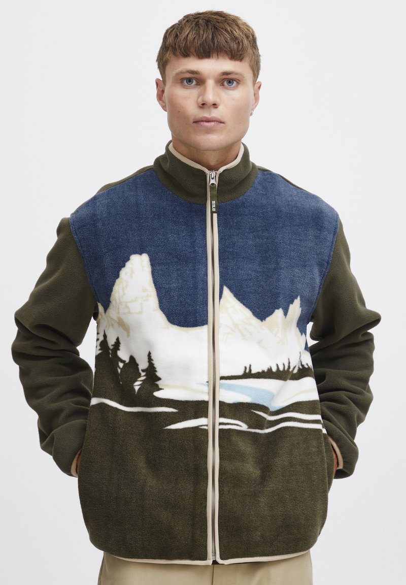Giacca in pile con colletto alto; design di un paesaggio montano blu e bianco sul davanti, maniche verde scuro e zip neutra.