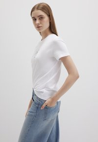 Weißes Baumwoll-T-Shirt mit kurzen Ärmeln, das ein geknotetes Design am Saum aufweist. Getragen mit hellblauen, lässig geschnittenen Jeans.