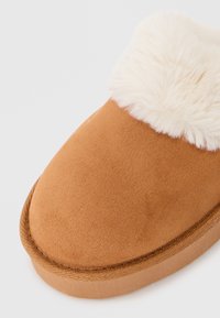 Pantofola in suede marrone con un risvolto in pelliccia sintetica bianca, punta arrotondata e suola piatta beige. Presenta una cucitura lungo il bordo.