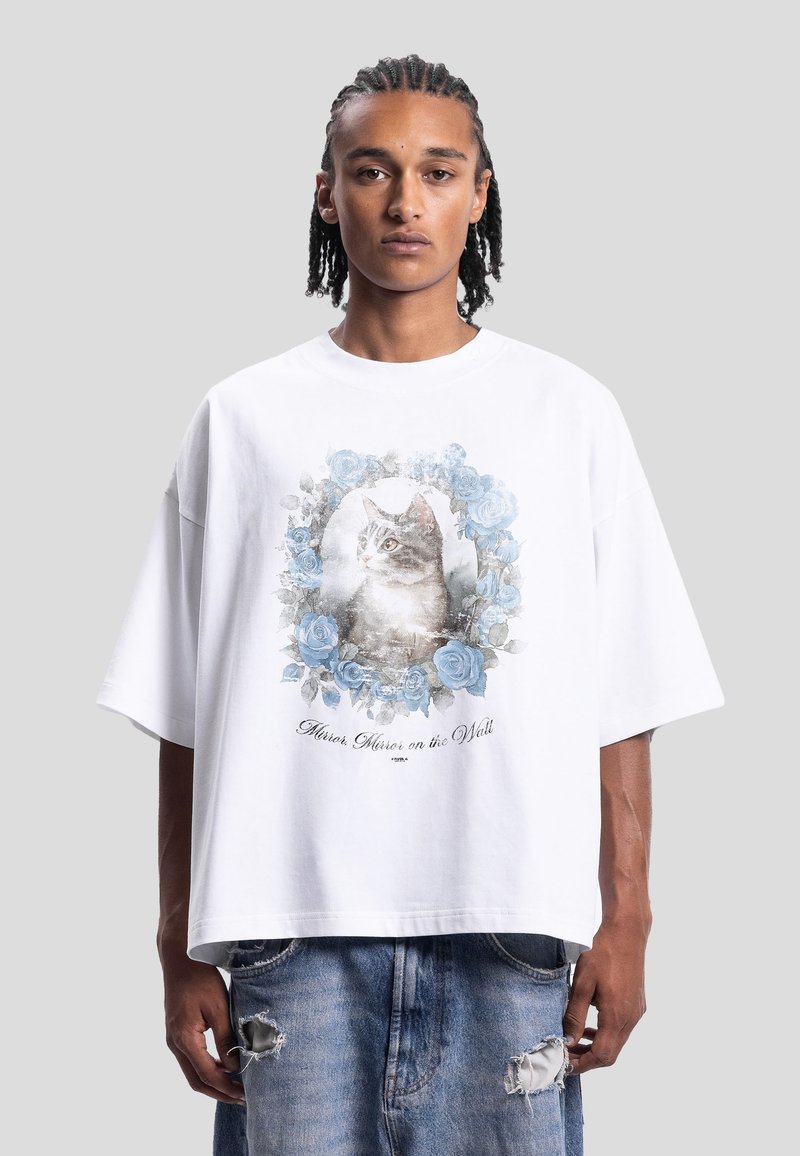 Homme portant un T-shirt blanc oversize avec un portrait de chat entouré de roses bleues et le texte « Miroir, miroir sur le mur. »