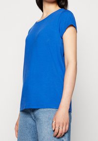 Modrá krátkosrstá tričko v uvolněném střihu, vyrobené z měkké tkaniny. Má kulatý výstřih a hladkou texturu. Nosené s světle modrými džíny.