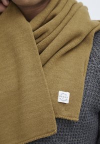 Strikket tørklæde i sennepsgul med et struktureret mønster, der har et hvidt mærke, der står "DENIM BLEND med ATTITUDE."