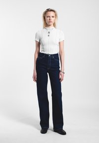 Bílý žebrovaný tričko s krátkými rukávy a vertikálními švy, spárované s tmavými denimovými zvonovými džíny s vysokým pasem a černými botami.