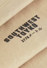 Vaatehkeet, joissa on kohotettua mustaa tekstiä, jossa lukee "SOUTHWEST TOYKO" ja "3776 メートル." Kankaassa on sileä ja kudottu rakenne.