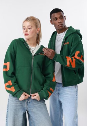 Ένας άνδρας και μια γυναίκα φοράνε πράσινες φούτερ με φερμουάρ και πορτοκαλί γράμματα, καθώς και ανοιχτόχρωμα τζιν, και στέκονται μπροστά σε λευκό φόντο.