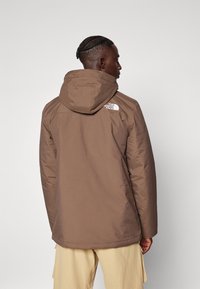 Brun isolert jakke med hette, med en hvit logo på øvre rygg og glatt tekstur, vist fra baksiden.