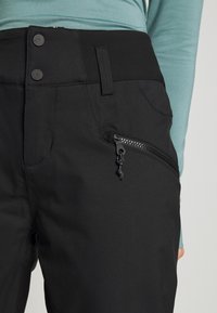 Pantalon noir à taille haute, doté de deux boutons-pression, d'une boucle pour ceinture et d'une poche latérale zippée, porté avec un haut à manches longues bleu sarcelle.