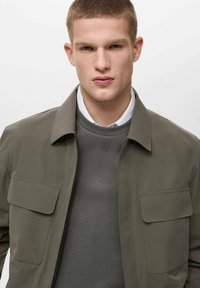 Olivgrüne Jacke mit zwei Brusttaschen, Reißverschluss und grauem Pullover darunter. Glattes Material, mit Kragen, strukturierte Form.