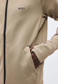 Personne portant une veste beige zippée avec le logo "BOSS", main dans la poche avant, détails visibles des coutures sur la manche et le côté.