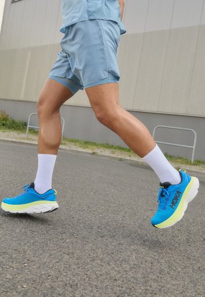 Personne courant sur l'asphalte portant des chaussures de course bleues, des chaussettes blanches et un short bleu clair devant un mur industriel gris.