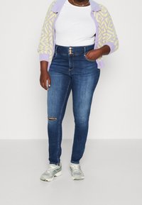 Vaqueros pitillo de tiro alto en azul oscuro con un detalle de rodilla rota, combinados con una camiseta blanca acanalada y un cárdigan estampado en púrpura y amarillo.