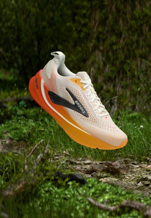 Lyse beige og orange Brooks løbesko med sort logo, vist flydende over skovbunden med grønne planter og sten.