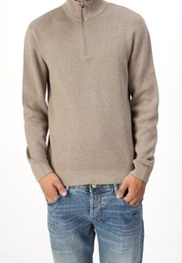 Beige zip-nek sweater met lange mouwen, geribbelde manchetten en zoom. Draag over lichtblauwe versleten spijkerbroek met een knoop en zakken.