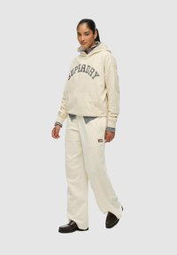 Vrouw in een crèmekleurige Superdry hoodie met een kangoeroezak, crèmekleurige wijde broek, gestreepte shirt eronder en donkerbruine schoenen, staand.