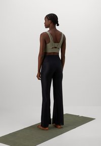 Sieviete stāv plika kāja uz zaļu jogas paklāja, ģērbusies olīvzaļā sporta krūšturī un melnās platās biksēs, pagriezusies prom no vienkāršas fona.