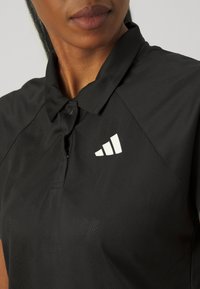 Mustolge T-särk, mis on valmistatud kergest kangast, millel on kolme nupuga krae ja valge adidas'i logo vasakul küljel.
