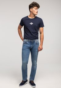 Mężczyzna w granatowej koszulce z logo, w wyblakłych niebieskich dżinsach i granatowych trampkach, stojąc i patrząc w lewo na jednolitym tle.