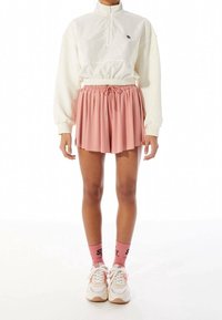 Witte ritshoodie met elastische taille, roze plissé rok, roze sokken met zwarte tekst en witte sneakers met perzikkleurige accenten.