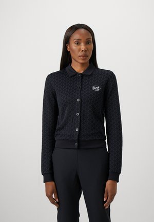 Vrouw in een zwart knopenjack met een subtiel ruitpatroon en het EA7-logo op de borst, gecombineerd met zwarte broek.