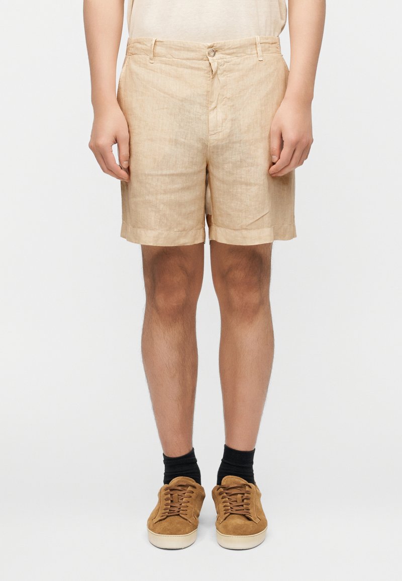 Ανοιχτό καφέ λινό σορτς με χαλαρή εφαρμογή, γυρισμένα τελειώματα και εμπρόσθιες τσέπες. Συνδυασμένο με καφέ σουέντ αθλητικά παπούτσια και μαύρες κάλτσες μέσης.