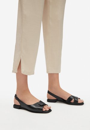Schwarze Ledersandalen mit offenem Zehendesign, verziert mit einer Schleife und verstellbarem Fersenriemen. Getragen zu hellbeigen, gekürzten Hosen.