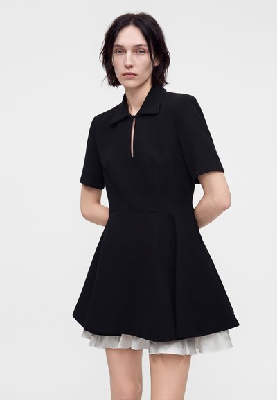 Femme aux cheveux courts et foncés portant une robe noire à manches courtes avec un col et un ourlet blanc volanté, debout devant un fond uni.