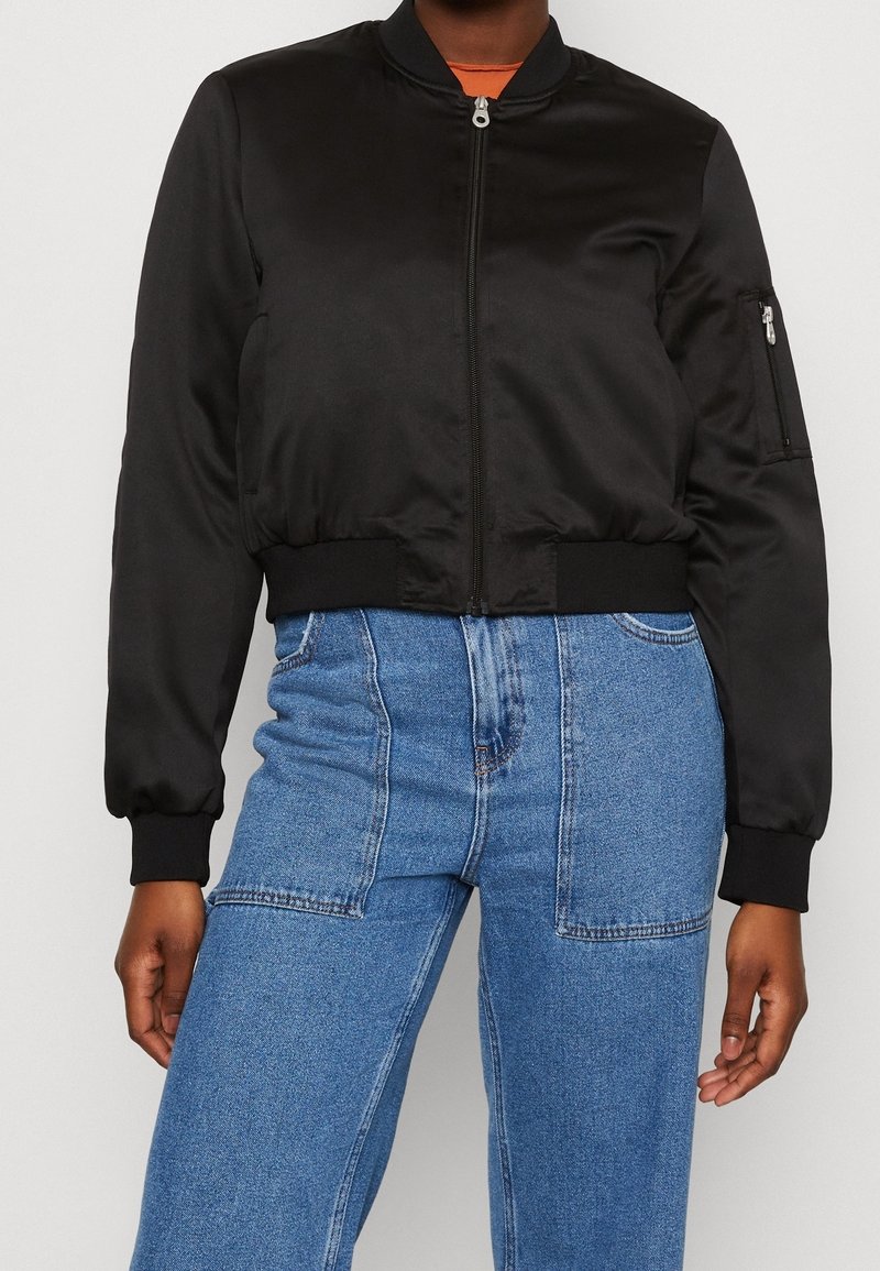 Personne portant une veste bomber noire zippée avec des poignets côtelés et un jean bleu taille haute avec de grandes poches avant.