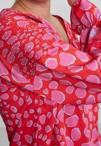 Blusa rossa e rosa con sezione superiore arricciata, dal taglio ampio, e con un motivo di macchie rosa di varie dimensioni sul tessuto.