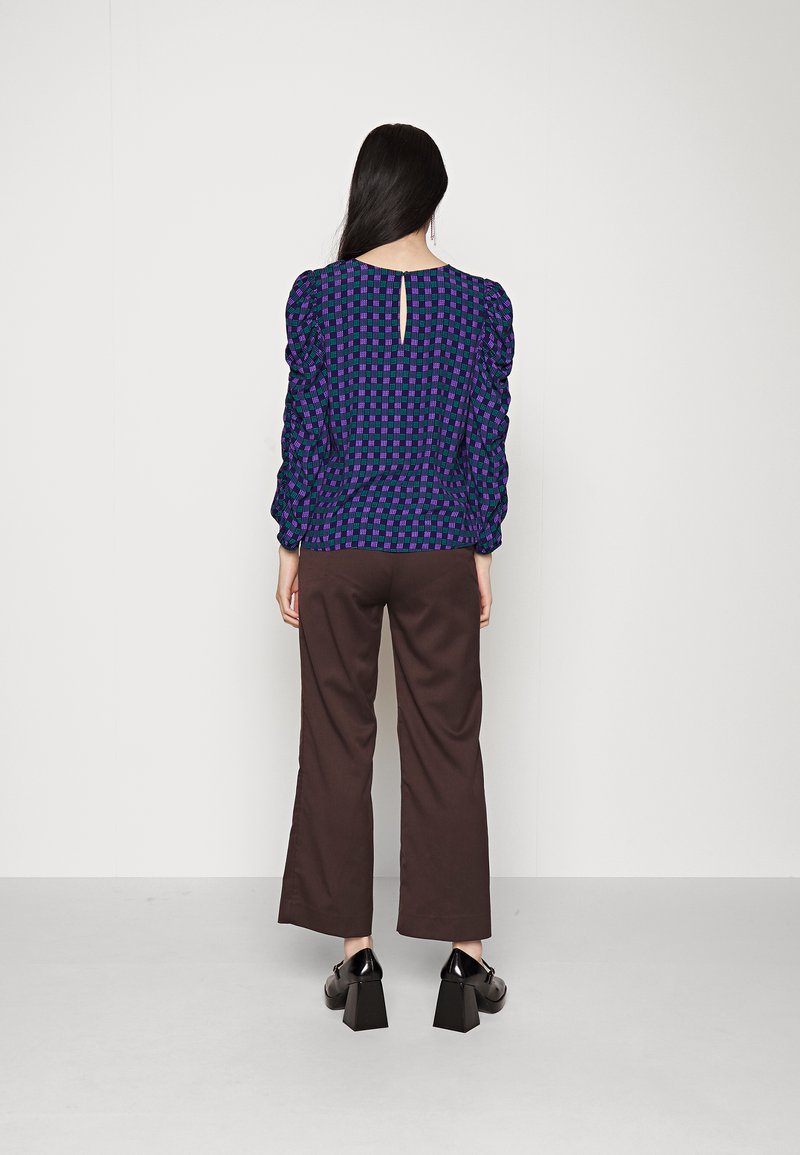 Blusa de manga longa em roxo escuro com padrão de xadrez verde; apresenta franzidos nos ombros e uma pequena abertura em forma de lágrima nas costas. Calças largas castanhas.