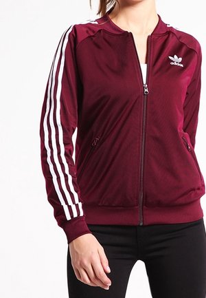 Vrouw draagt een bordeauxrode Adidas trainingsjack met witte drie strepen op de mouwen, dichtgeritst, gecombineerd met zwarte broek.