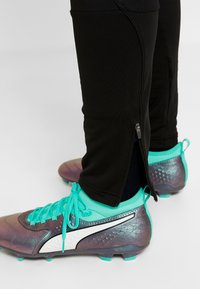 Zwarte sportbroeken met ritsdetails bij de enkels, gecombineerd met teal en grijze voetbalschoenen met een gestructureerd ontwerp en veters.