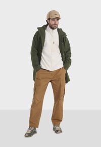 Grüne wasserdichte Jacke über einem weißen Fleecepullover, kombiniert mit beige Cargo-Hosen und grau-grünen Sneakers. Merkmale sind eine Reißverschlusstasche und verstellbare Elemente.