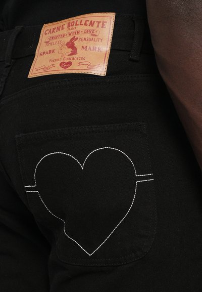 Svarta denimjeans med en brun läderlapp som har "Carne Bollente"-logotyper. Vit hjärtformad söm på bakfickan.