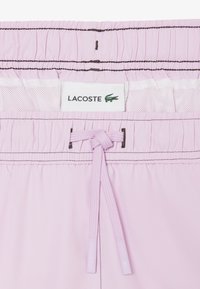 Shorts de bain rose clair avec ceinture élastique, cordon de serrage noué, doublure en maille blanche et étiquette logo Lacoste avec crocodile vert.