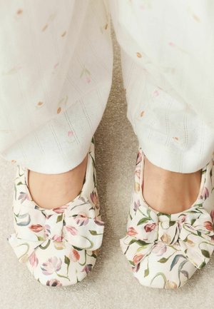 Füße, die weiße, mit Blumen verzierte Hausschuhe mit großen Schleifen tragen, stehend auf einem beigen Teppich, kombiniert mit weißen Hosen mit dezenten rosa und orangefarbenen Mustern.
