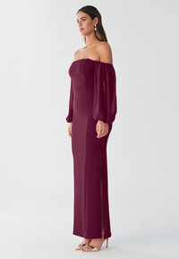 Burgundska obleka z odkritimi rameni, dolgimi prosojnimi rokavi, silhueto, ki objema telo, stranskim razporkom in gladkim materialom. Kombinirana z metalik petkami.