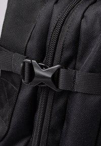 Black backpack with textured nylon material. Features a zipper closure and plastic buckle. The design is compact with reinforced stitching.