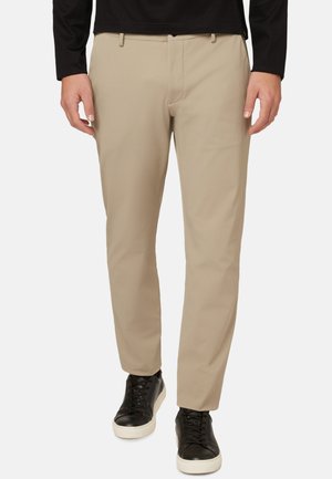 Pantalons beige ajustés en tissu lisse avec une taille plate, associés à des baskets noires avec une semelle et des lacets blancs.