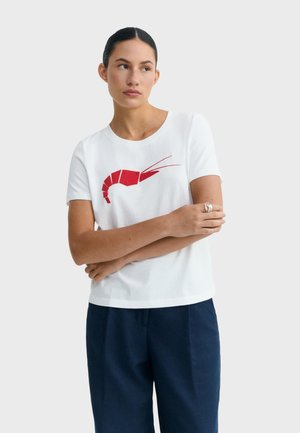 Woman with dark hair in a bun wears a white t-shirt with a red shrimp graphic, dark blue pants, and a silver ring, posing with arms crossed.